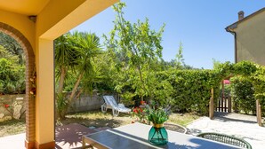 Une terrasse avec une table, des chaises, un vase de fleurs et une vue sur un jardin agrémenté d’un mur de briques et d’une porte en bois.