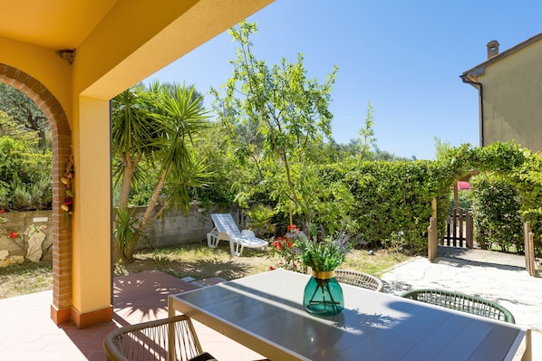 Um pátio com mesa, cadeiras, um vaso de flores e vista para um jardim com muro de tijolos e portão de madeira.