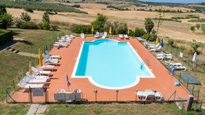 Une piscine rectangulaire, bordée d’une terrasse carrelée, entourée de chaises longues et de parasols, le tout se fondant dans un paysage de collines douces et sous un ciel dégagé.