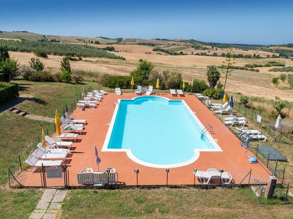 A rectangular swimming pool with a tiled deck, surrounded by lounge chairs and umbrellas, set against a backdrop of rolling hills and a clear sky.
