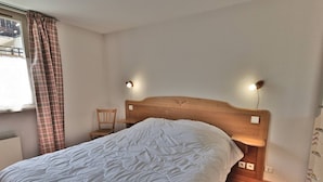 Un dormitorio con cabecera de madera, cubrecama blanco, dos lámparas de noche, una silla de madera y una ventana con cortinas a cuadros.