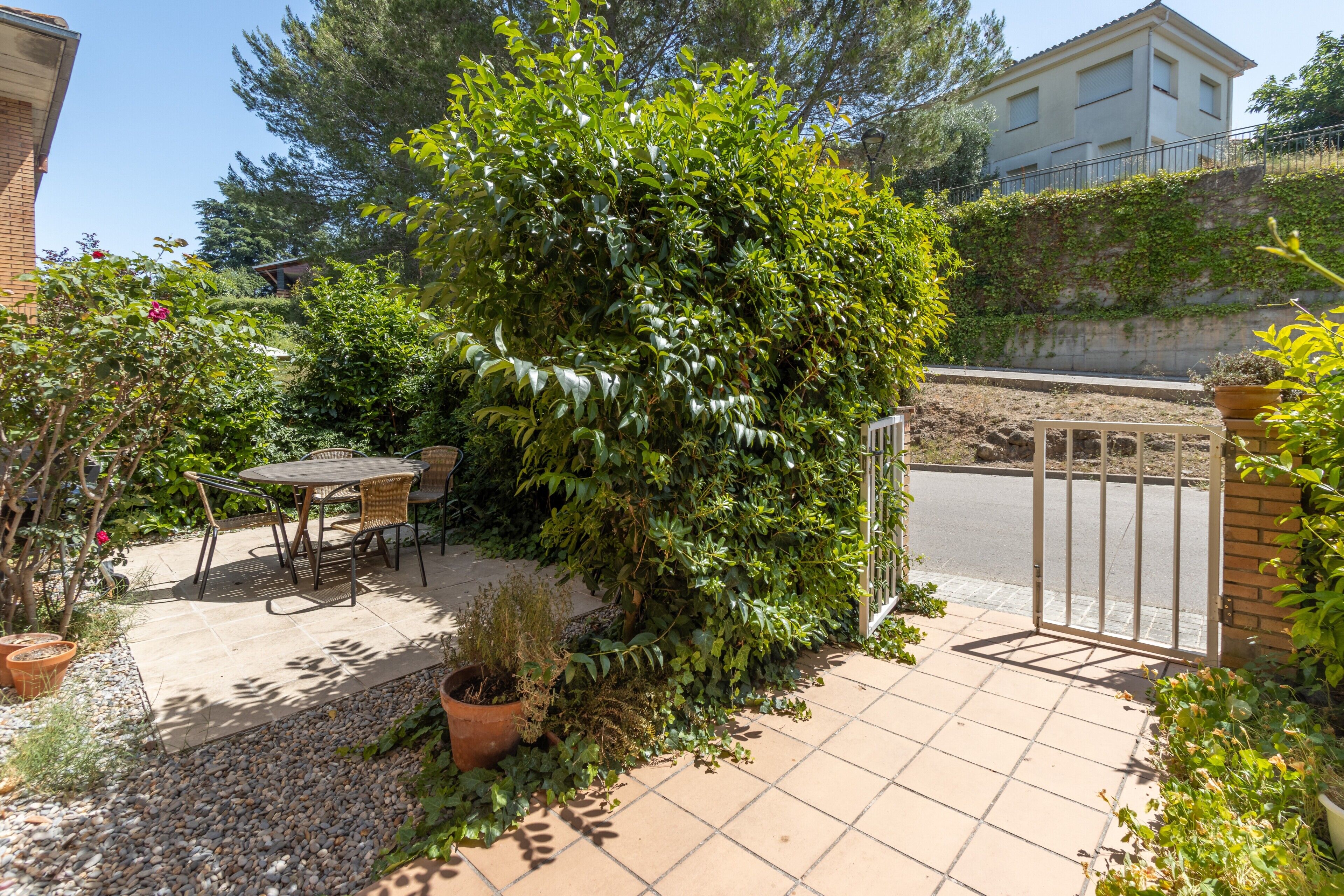 A patio with a table and chairs, surrounded by greenery and a view of a building in the background.