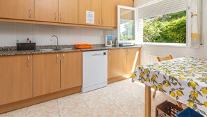 Een keuken met houten kastjes, een vaatwasser, een raam met zonwering en een tafel met een tafelkleed met citroenmotief.