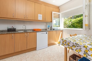 A kitchen with wooden cabinets, a dishwasher, a window with blinds, and a table with a lemon-patterned tablecloth.