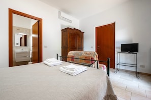 Un dormitorio con cama, armario, televisor sobre un mueble bajo y baño visible a través de una puerta abierta.