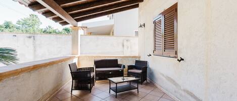 A covered patio with a wooden ceiling, a cushioned bench, two wicker chairs, a small table, and a wall-mounted light fixture.