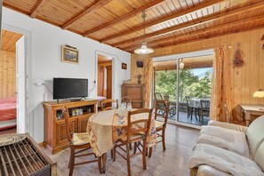 Ein Holzinterieur mit einem Esstisch, Stühlen, einer Couch, einem Fernseher und Blick durch eine Glastür.