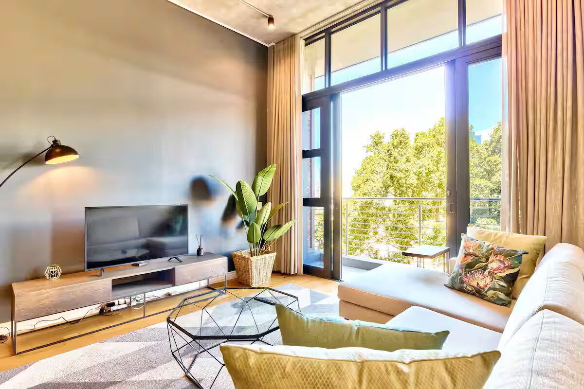 Een moderne woonkamer met een flatscreen televisie, een bank, een salontafel en een groot raam met uitzicht op groen.