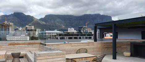 Terraço no telhado com piso de madeira, mobiliário e vista para paisagem urbana e montanhas.