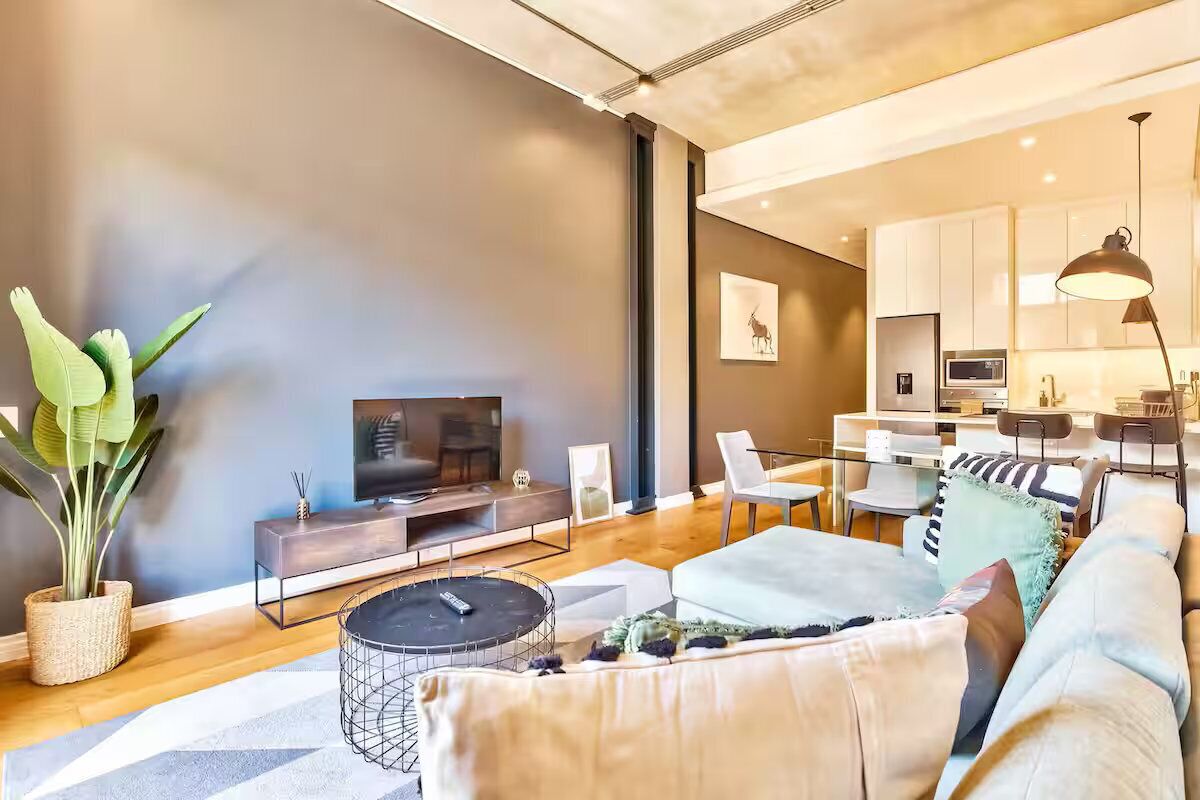 Una sala de estar moderna con un sofá, una mesa de centro y un televisor. Se observa un comedor con sillas y una mesa.