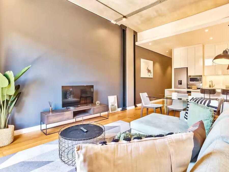 Una sala de estar moderna con un sofá, una mesa de centro y un televisor. Se observa un comedor con sillas y una mesa.