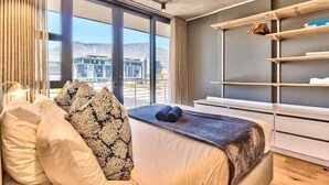 A modern hotel room with a large bed, a bedside table, a wardrobe, and a view of a cityscape through large windows.