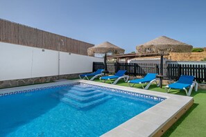 A swimming pool with blue tiles, surrounded by lounge chairs and thatched umbrellas.