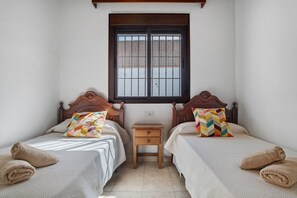 A room with two beds, a small wooden nightstand, a window, and a light switch on the wall.