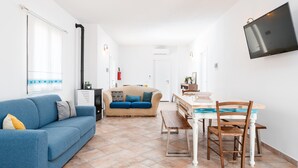 Een woonkamer met een blauwe bank, een beige bank, een eettafel met stoelen, een aan de muur bevestigde televisie, en een keuken in de achtergrond.