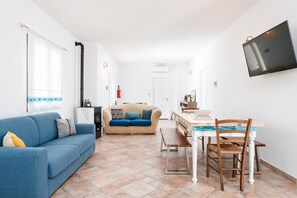 A living room with a blue sofa, a beige sofa, a dining table with chairs, a TV mounted on the wall, and a kitchen area in the background.