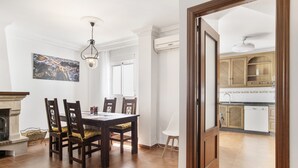 Un coin repas avec une table et des chaises en bois, une cheminée et une cuisine visible à travers une porte ouverte.