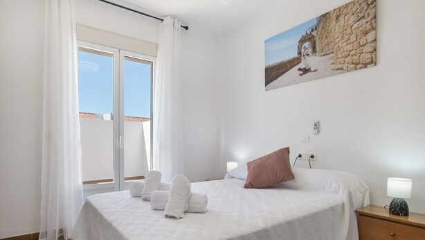 Une chambre à coucher avec un lit, une table de chevet, une lampe, un tableau et une fenêtre avec des rideaux.