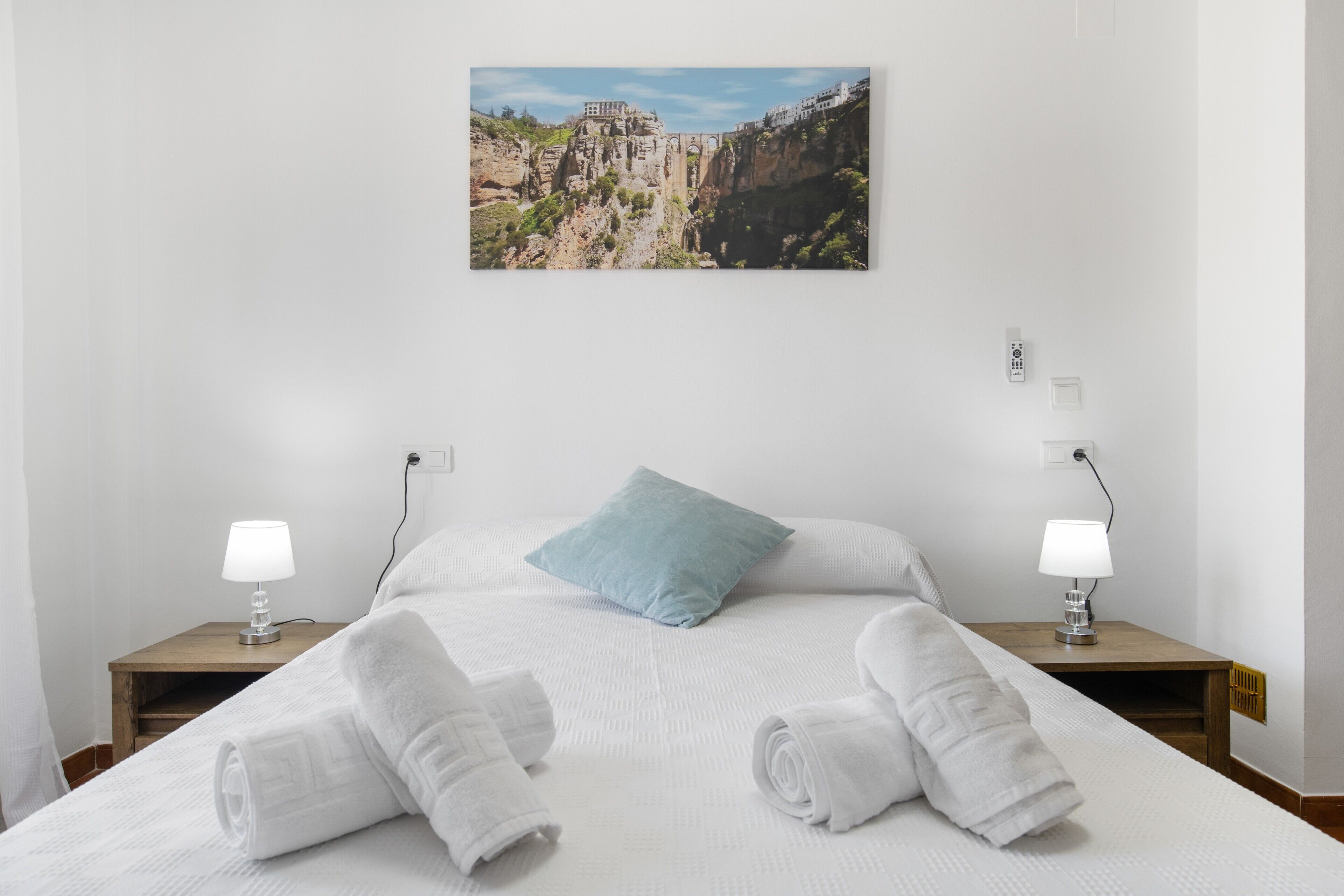 Une chambre d’hôtel avec un lit, deux lampes de chevet, un seul oreiller et un tableau représentant un paysage montagneux accroché au mur.