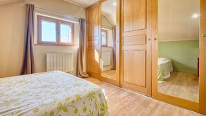 Un dormitorio con cama, armario, espejo, radiador y ventana con cortinas.