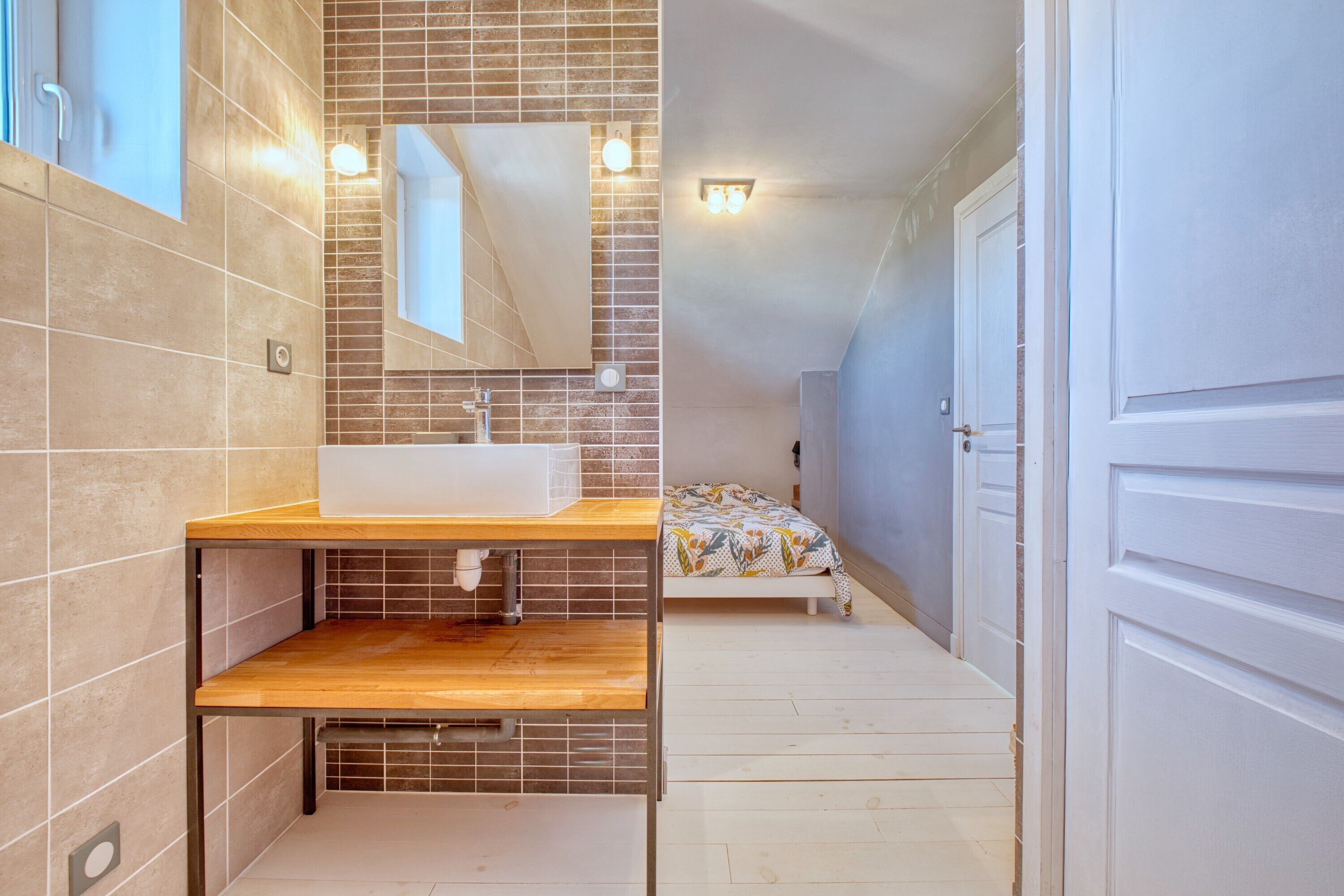 Une salle de bain avec un meuble-lavabo en bois, un grand miroir, des murs carrelés, un lit avec une couvre-lit à motifs et une porte.