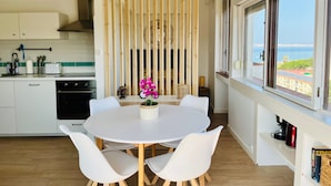 A modern kitchen with white cabinets, a wooden divider, a round dining table with white chairs, and a view of the sea through large windows. - Apartment 'Sunset' with Sea View, Wi-Fi and Air Conditioning (Alvor)