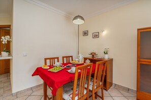 Uma sala de jantar com toalha de mesa vermelha, cadeiras de madeira, um candelabro e uma cozinha ao fundo.