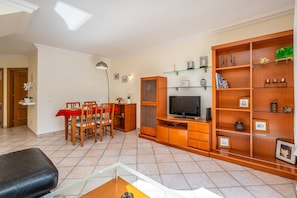 Un salon avec un téléviseur, des étagères en bois, une table à manger avec des chaises et une nappe rouge.