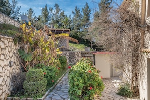 Un sentiero in pietra conduce a un edificio rustico con un muro in pietra, un piccolo deposito e vegetazione rigogliosa.