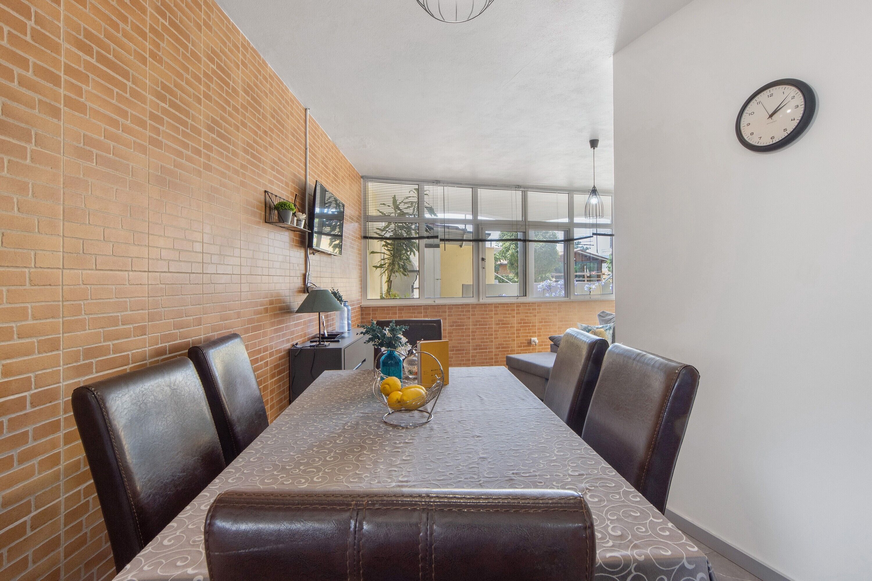 A dining area with a brick wall, a table with a patterned tablecloth, and a clock on the wall.