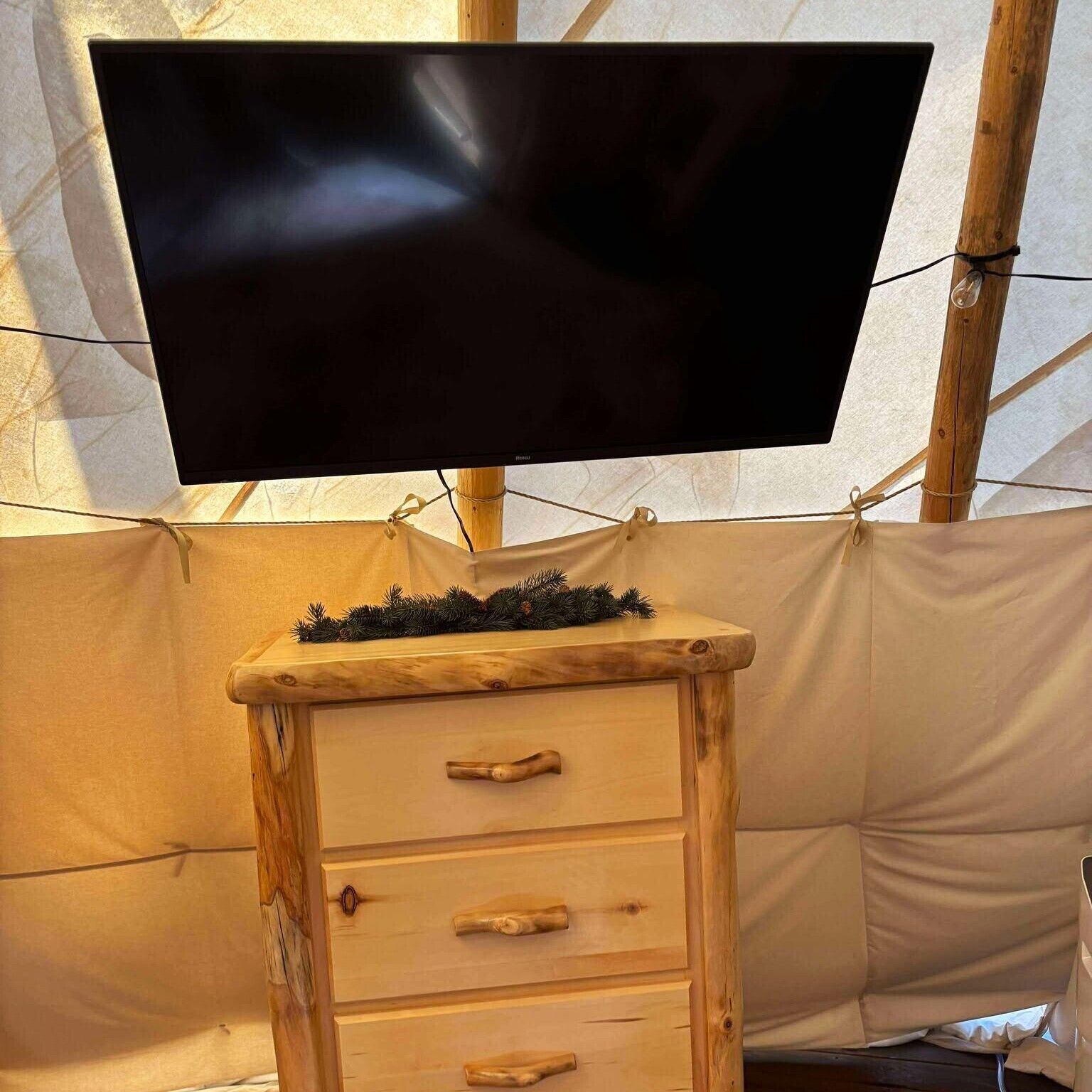 A television mounted on a wooden stand with a decorative pine branch, set against a backdrop of a tent-like structure.