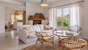 A spacious living area with a white sofa, a round wooden table, and wicker chairs. A fireplace with woven baskets is visible, and the room has large windows with curtains. - Villa Punta – 2/3 Bedrooms, Pool, Domaine de la Pointe (Grosseto-Prugna)