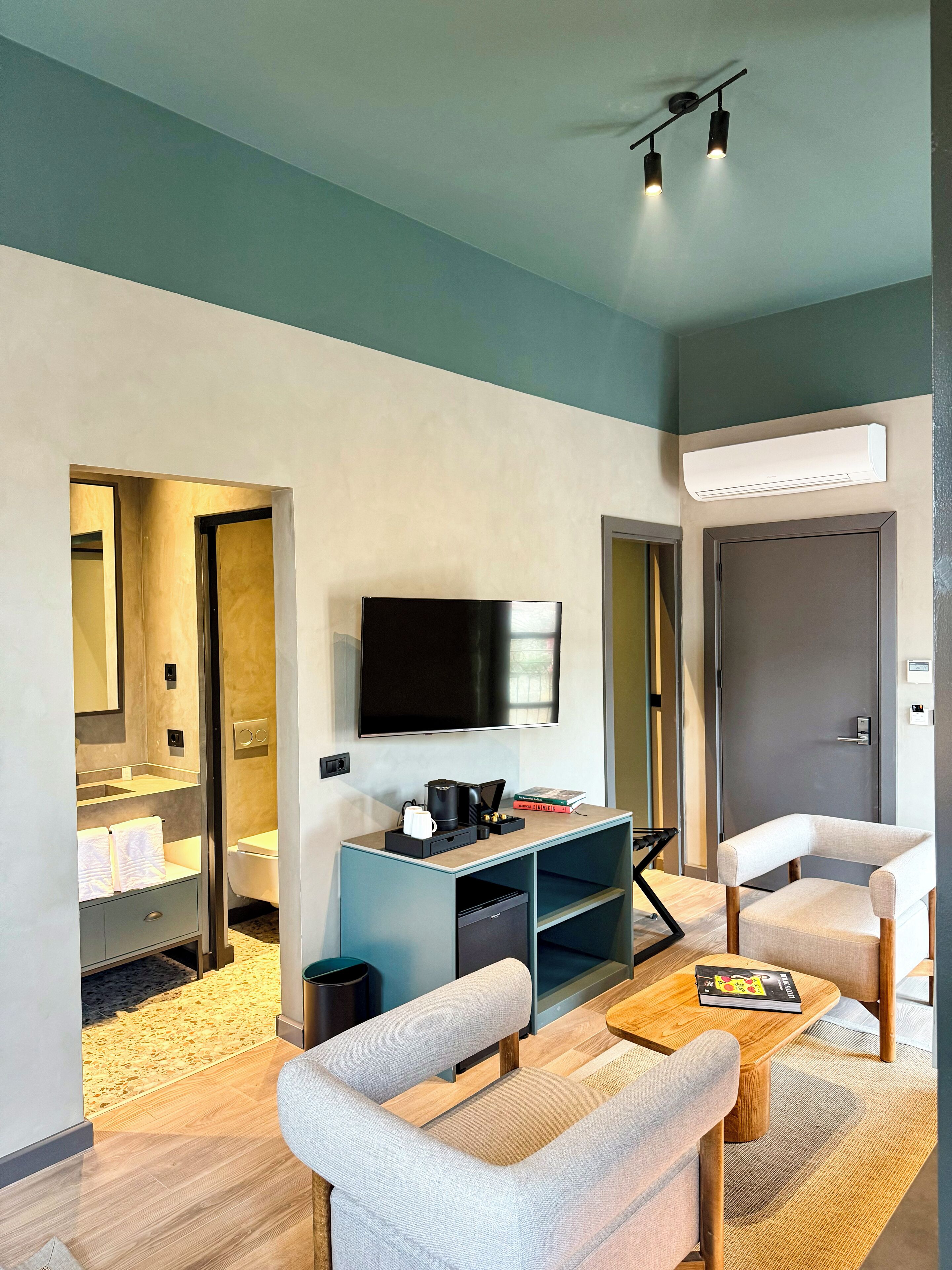 Ein modernes Hotelzimmer mit Flachbildfernseher, einer Kaffeemaschine, einem Bücherregal und einer Sitzecke mit zwei Stühlen und einem kleinen Tisch.