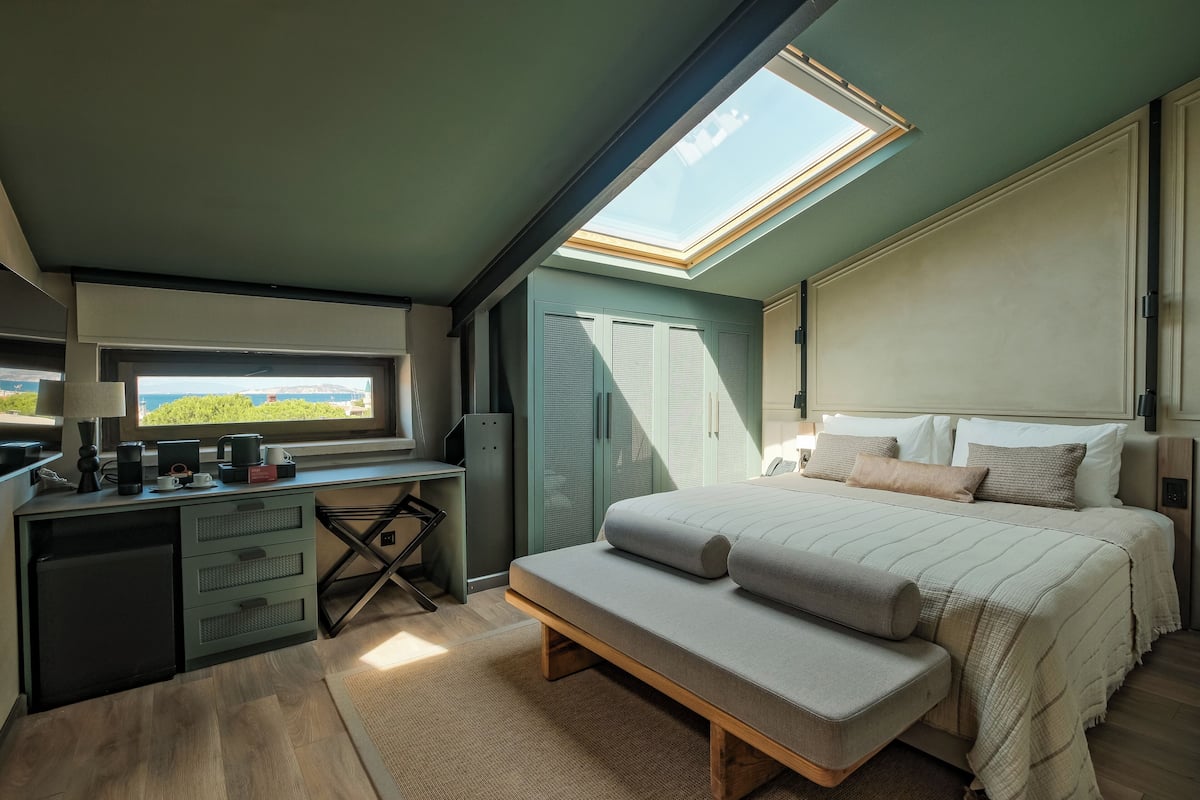 Un dormitorio con tragaluz, una cama con banqueta, un escritorio con computadora y vista a la vegetación exterior.