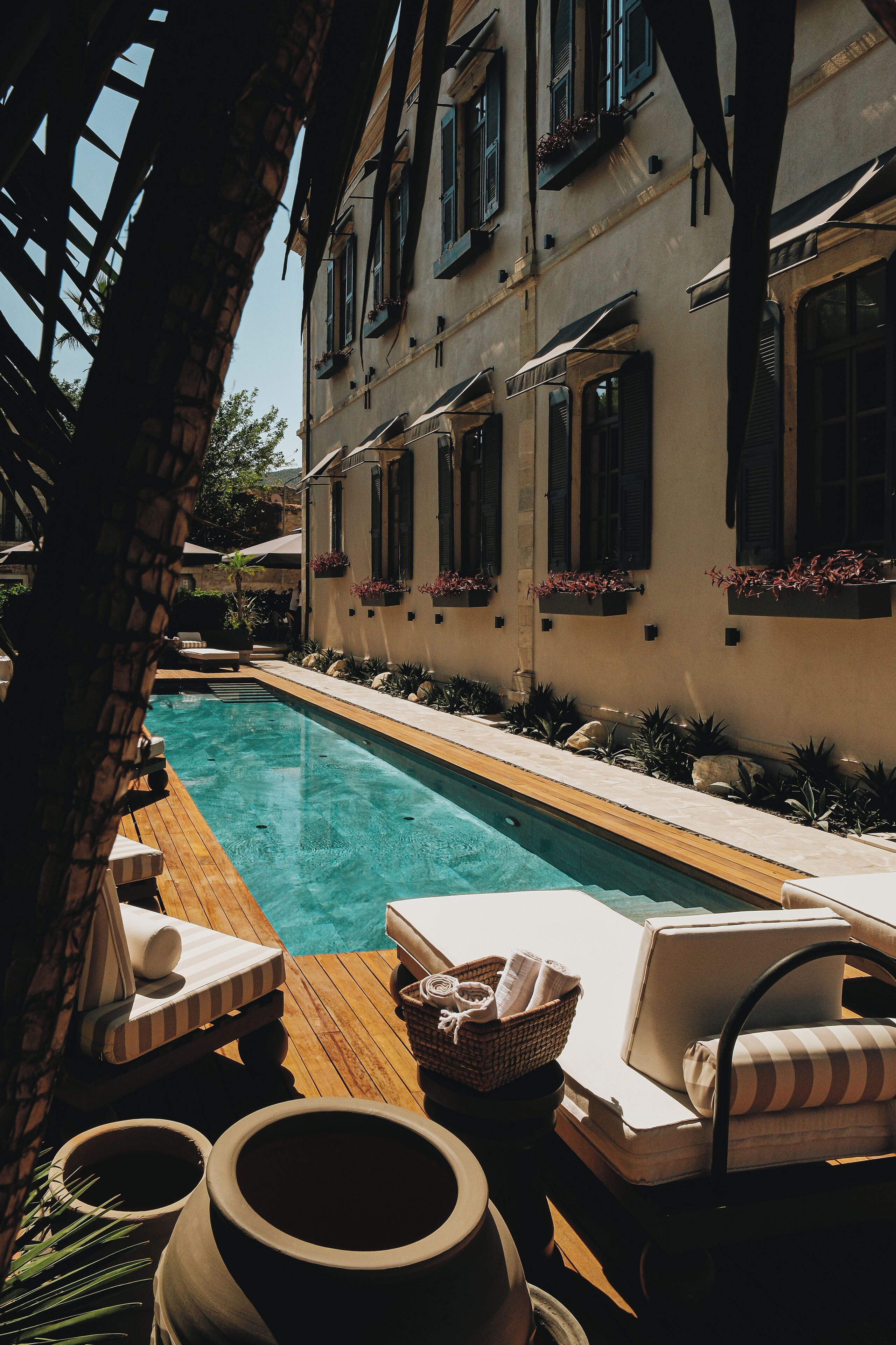 Un espace aménagé au bord de la piscine, avec des chaises longues, une terrasse en bois et un bâtiment doté de fenêtres et de jardinières.