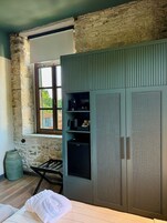 Una habitación con pared de piedra, ventana, cama, armario y una pequeña mesa con silla.