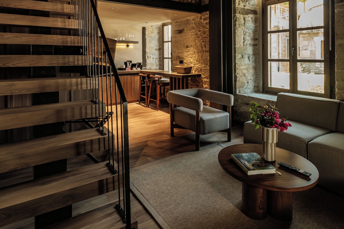 A spacious living area with a wooden staircase, a sofa, a coffee table, and a view of the city through large windows.