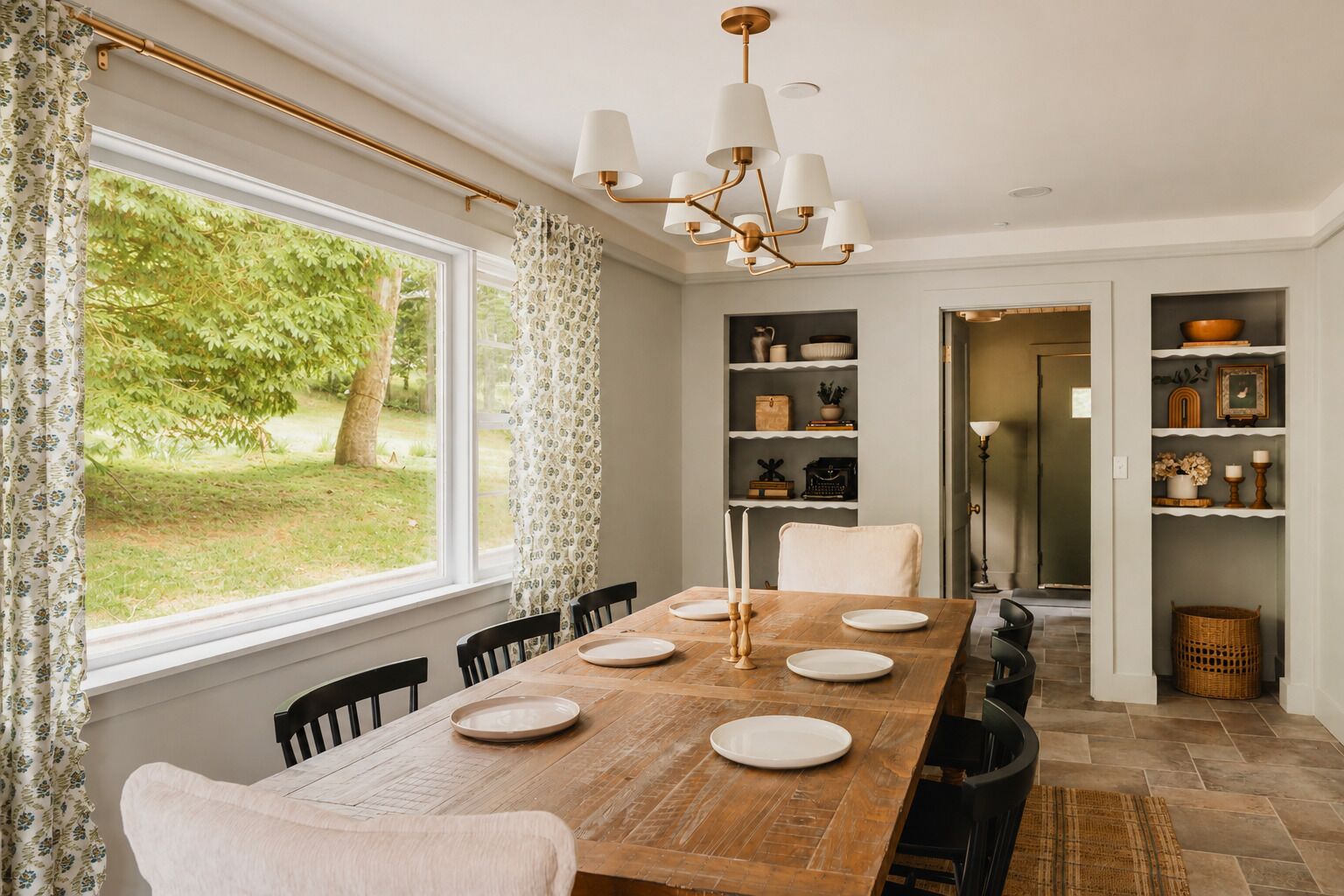 A dining room with a wooden table, chairs, and a chandelier. There are shelves with decorative items and a view of greenery outside.