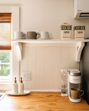 A kitchen with a white countertop, a coffee maker, sugar and coffee containers, and a window with a view of greenery.