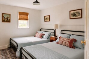 A room with two single beds, a window with blinds, a bedside table with a lamp, and framed artwork on the walls. - Hot Tub, Fire Pit, Porch | 1850s Riverside Cabin (Barryville)
