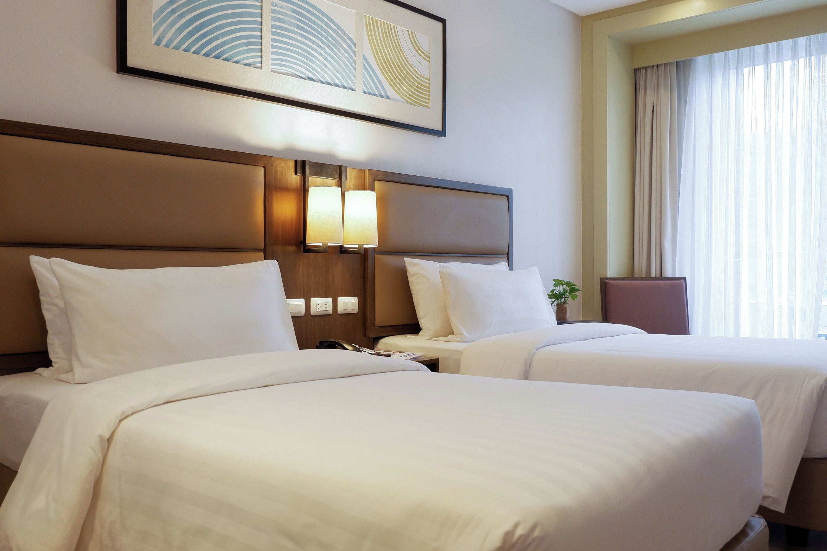 Habitación de hotel con dos camas, cabecera con iluminación integrada, ventana grande con cortinas translúcidas y una obra de arte abstracta enmarcada sobre las camas.