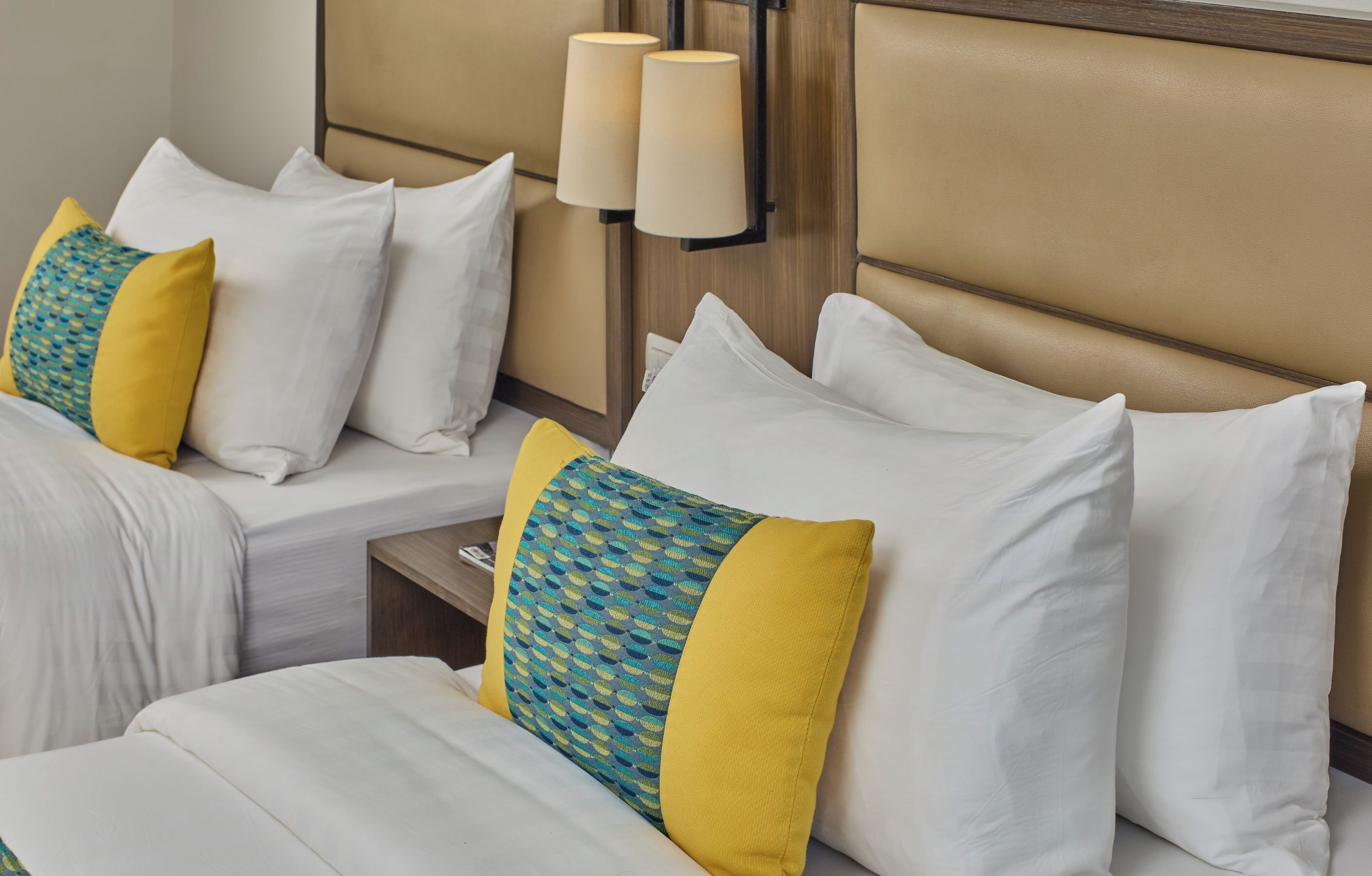 Une chambre d’hôtel avec un lit, du linge de lit blanc et des coussins décoratifs. On y trouve deux lampes fixées au mur et une tête de lit en bois.