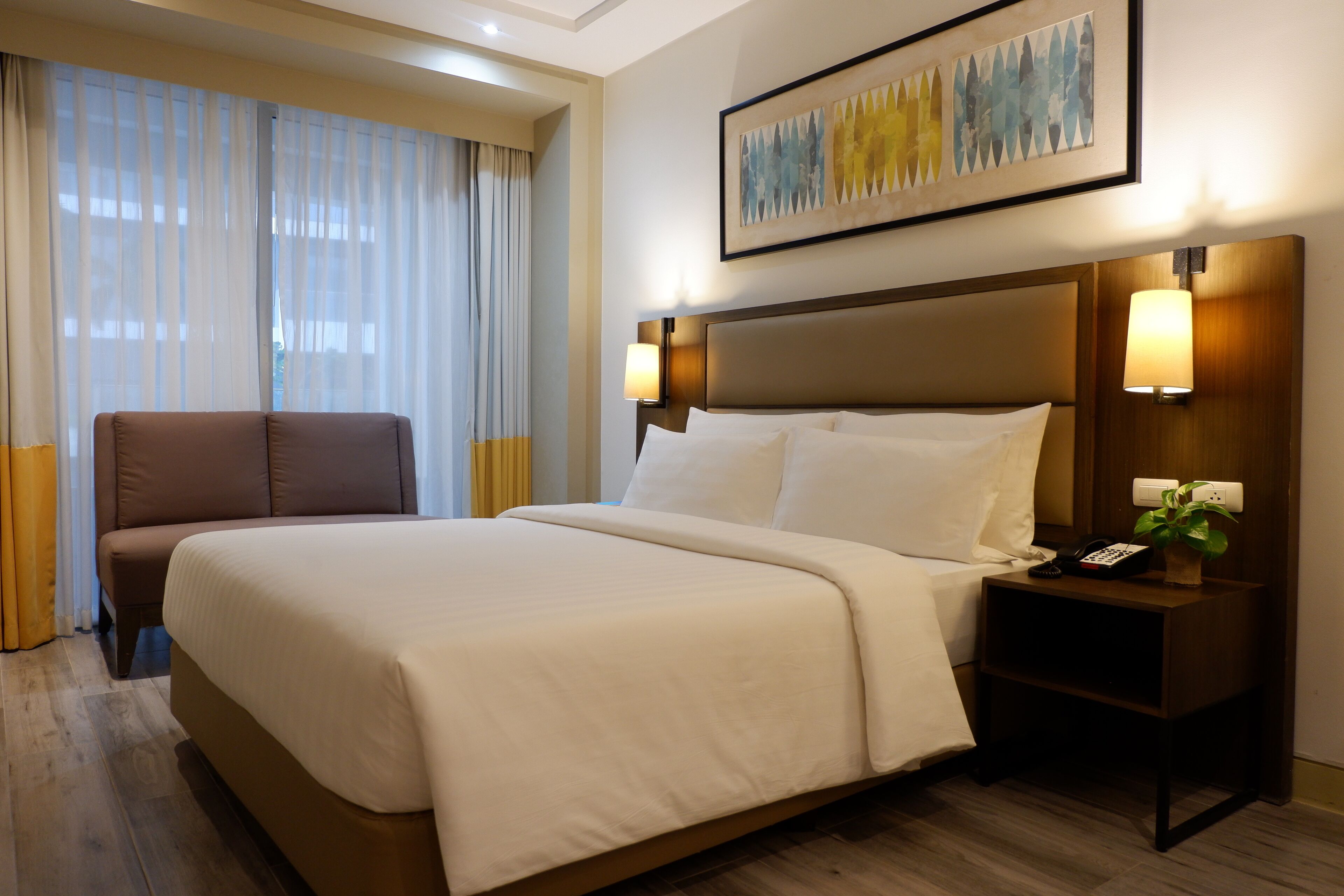 Una habitación de hotel con una cama grande, una mesita de noche con lámpara, una silla y un cuadro en la pared.