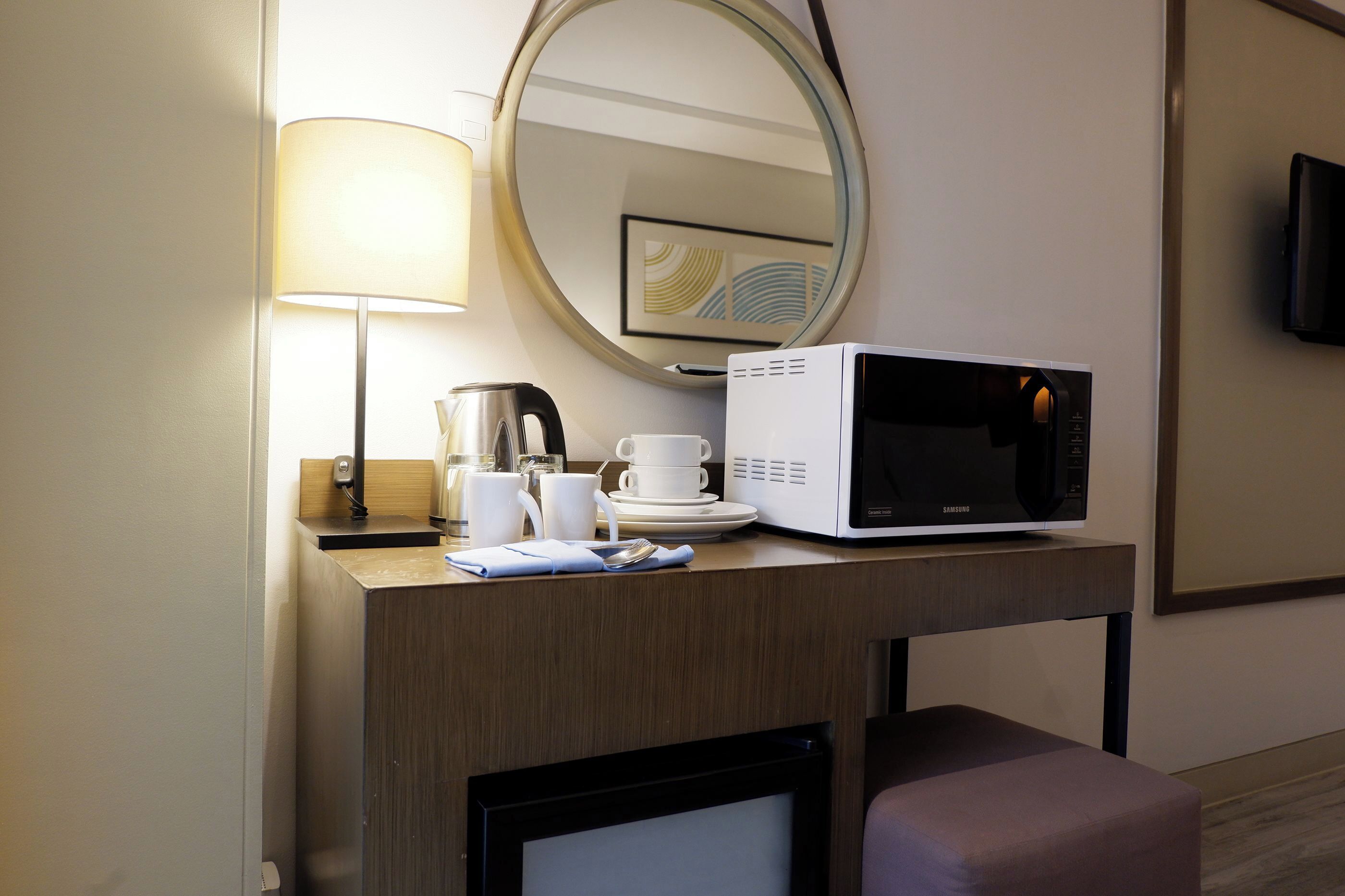 A hotel room with a wooden desk, a microwave, a coffee maker, a lamp, and a television on the wall.