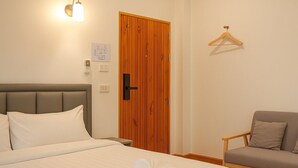 Un lit bien fait, avec du linge de lit blanc et deux serviettes posées dessus. On y voit une porte en bois avec une poignée, un interrupteur et un luminaire fixé au mur. Une chaise en bois avec des coussins est placée contre le mur.
