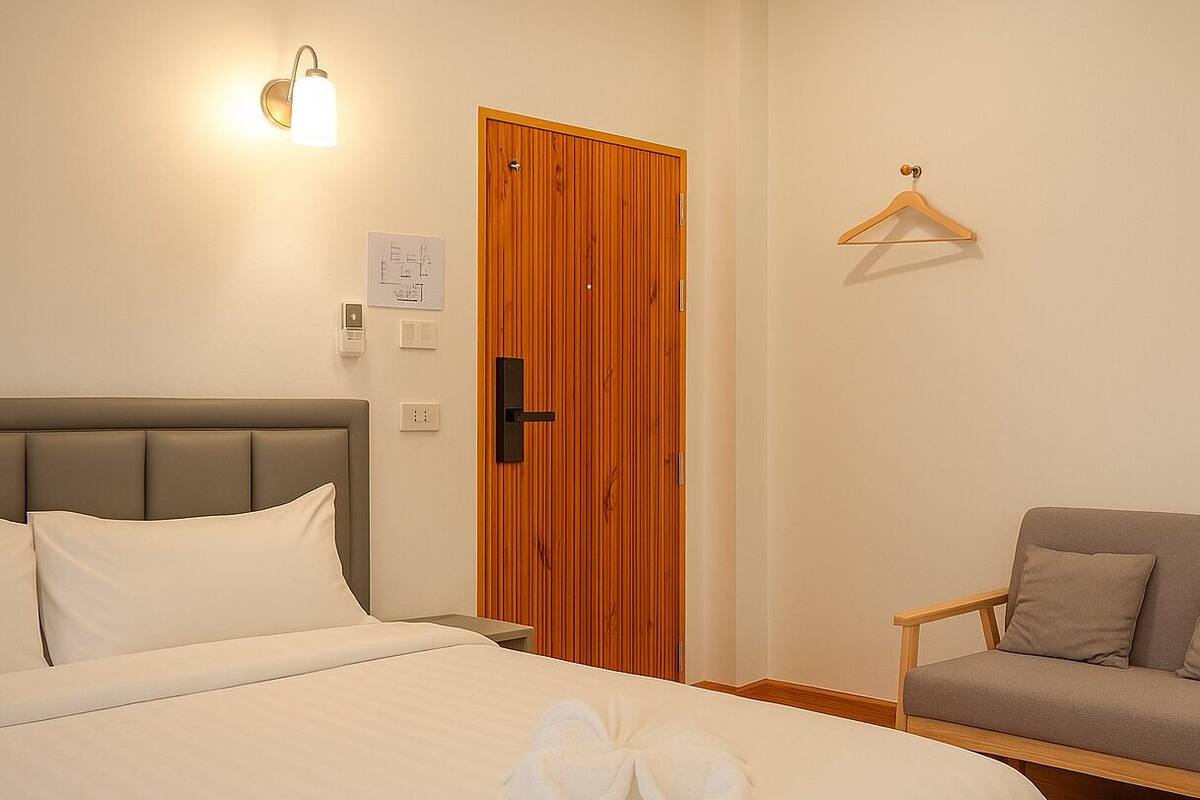 Een net opgemaakt bed met witte lakens en twee handdoeken erop. Er is een houten deur met een handvat, een lichtschakelaar en een wandlamp. Een houten stoel met kussens staat tegen de muur.
