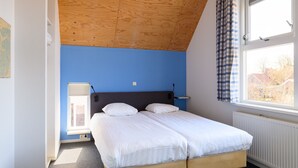 Un dormitorio con techo inclinado, cama con ropa blanca, ventana con cortinas a cuadros y un radiador.