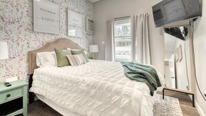 Una camera da letto con carta da parati a motivi floreali, un letto con biancheria bianca, due quadri appesi, una televisione e uno specchio.