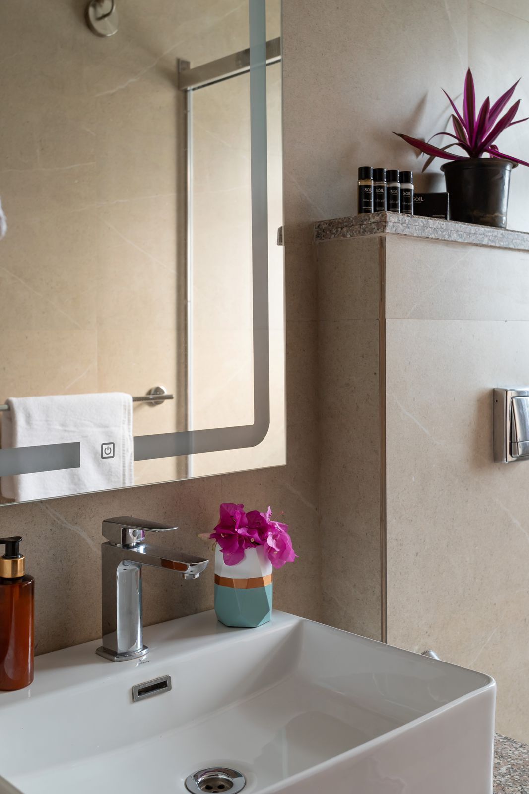 Un baño con lavamanos blanco, un espejo, toallero, planta en maceta y artículos de aseo en un estante.