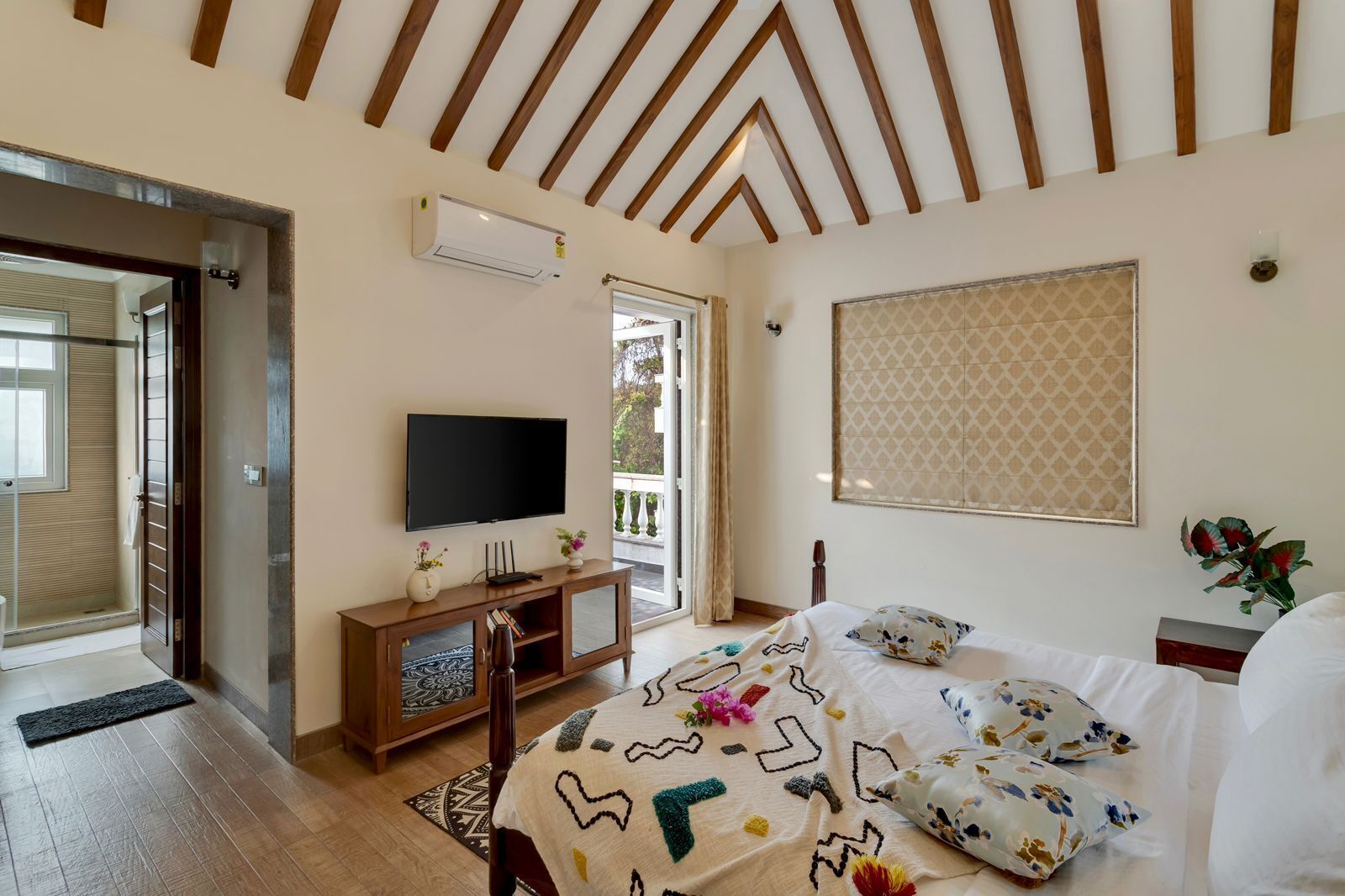 A bedroom with a TV, wooden furniture, a patterned bedspread, and a window with a view.